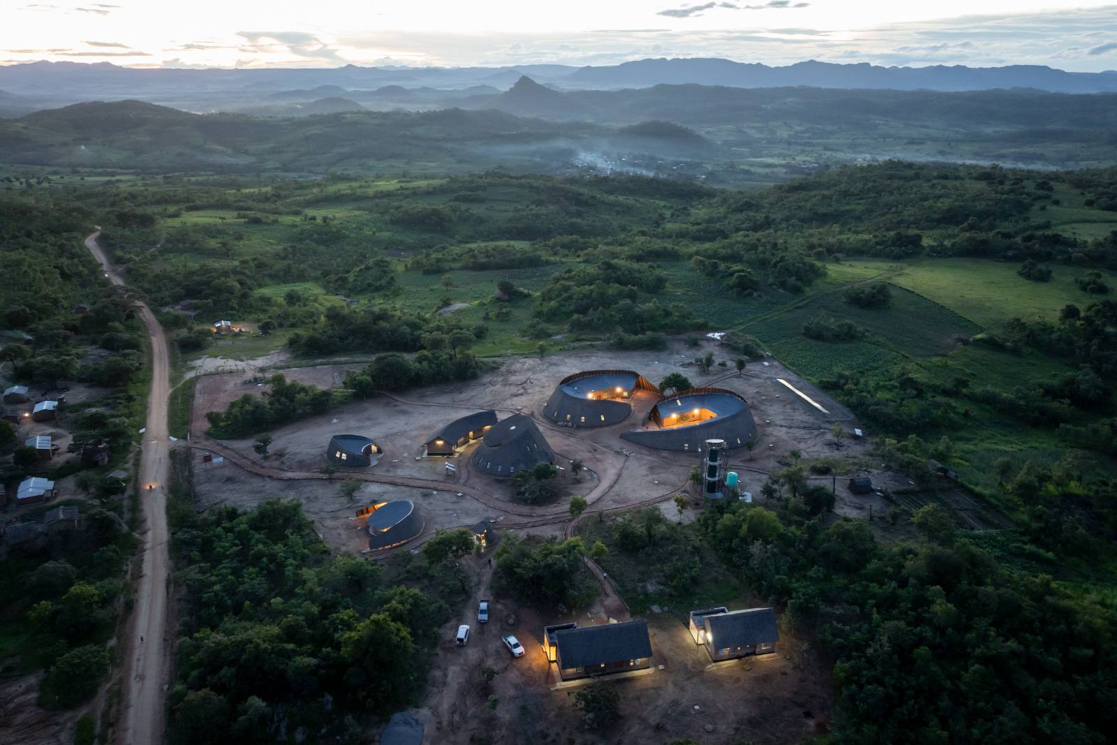 Aerial view of the campus at dusk with interior lights on, the road running past and hills receding to the horizon