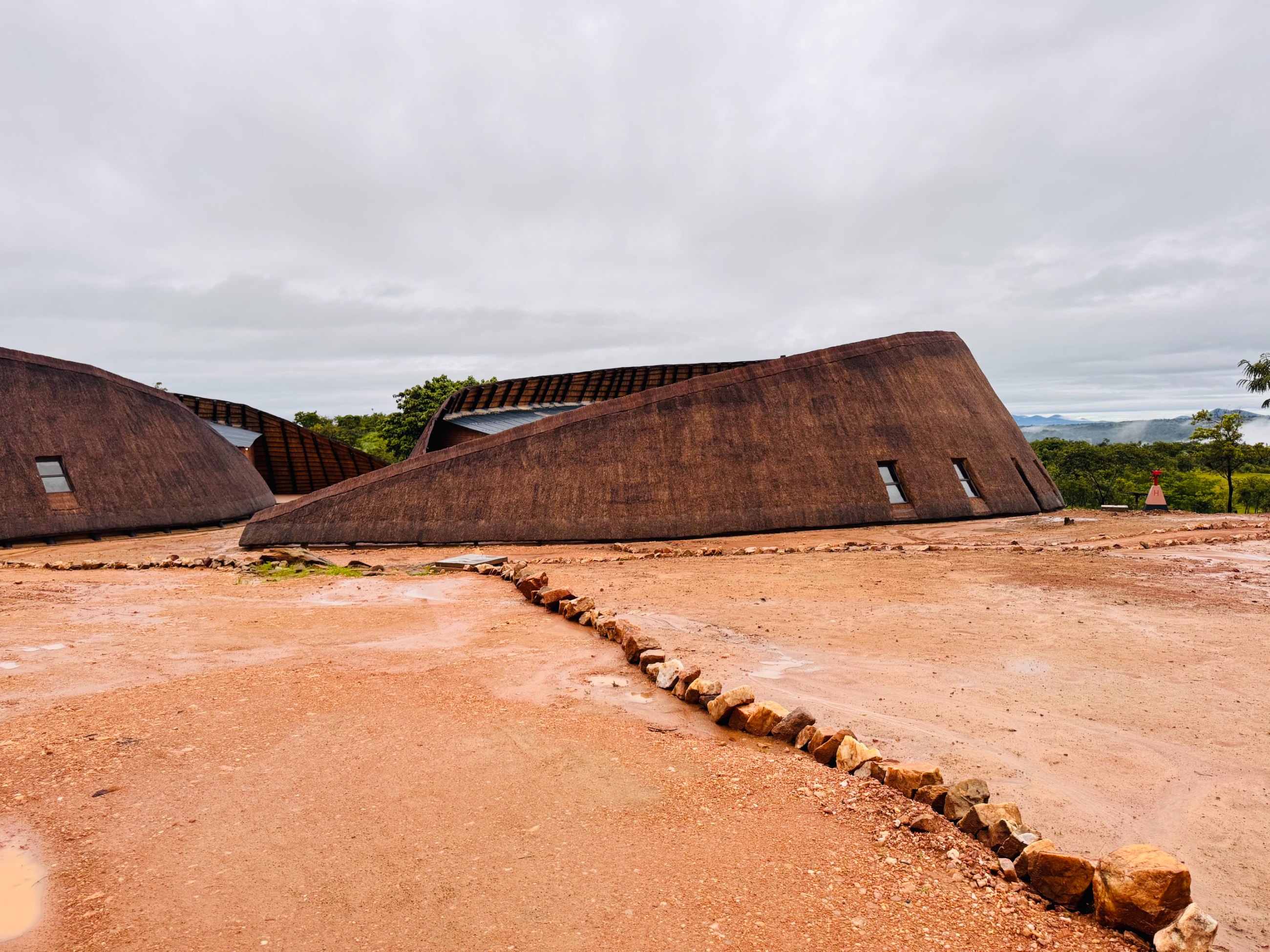 Two thatched halls with long sweeping roofs meeting the red earth under an overcast sky, hills in the distance