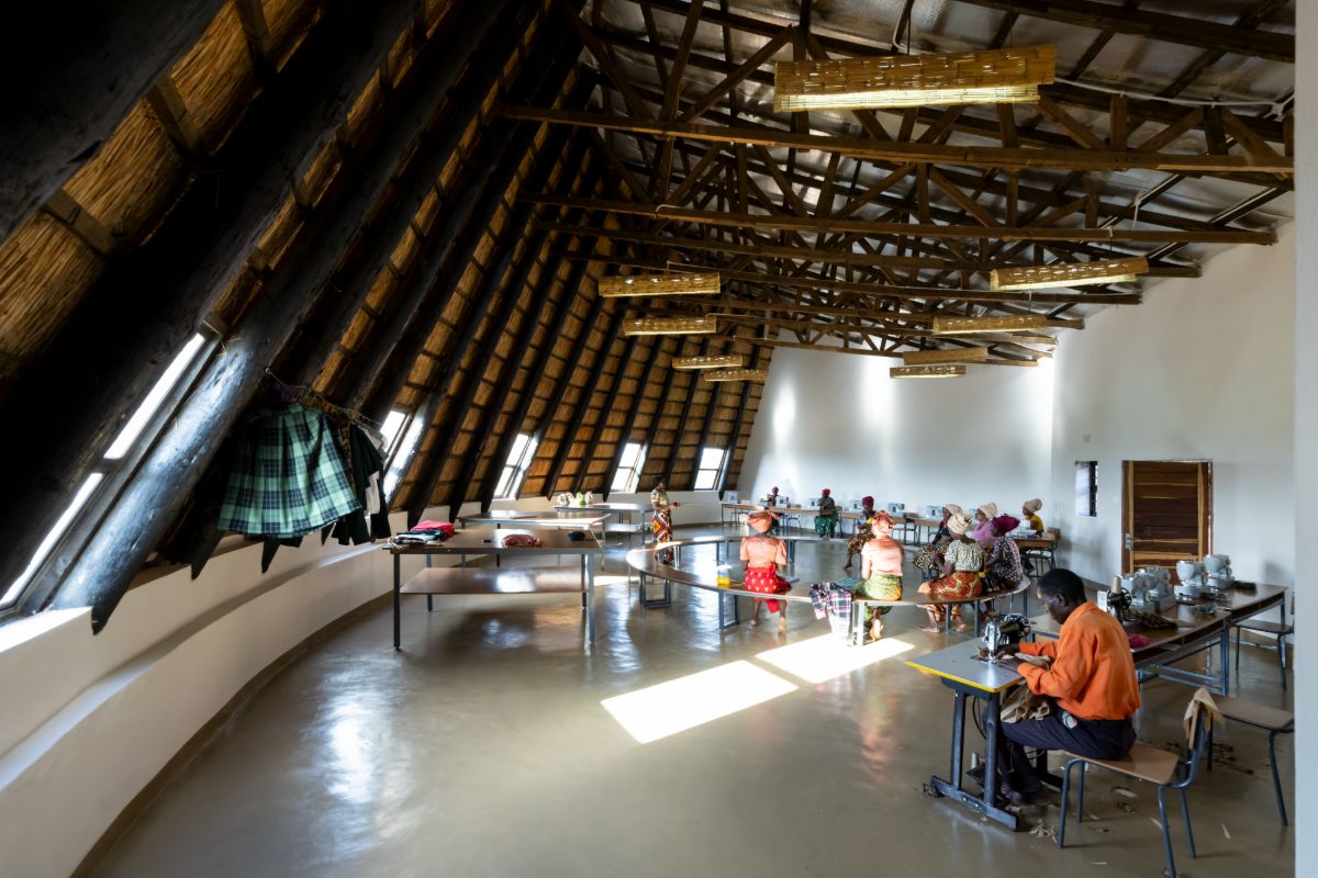 A tailoring workshop under a thatched roof on timber trusses, people at sewing machines along the tables and a group seated at a curved bench